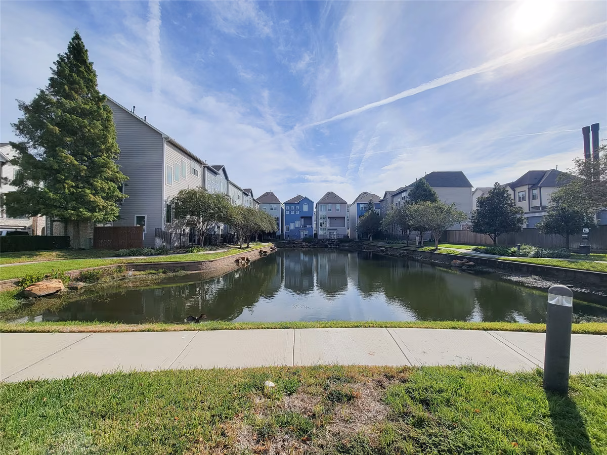 Charming neighborhood view featuring lining a serene pond, complemented by lush greenery and a clear blue sky.