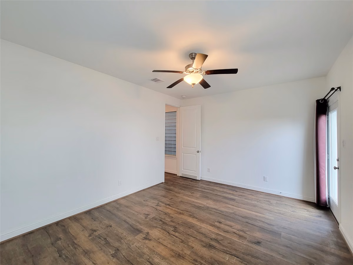 Spacious room with elegant wood flooring, a modern ceiling fan, and ample natural light from a glass door, perfect for a cozy retreat.